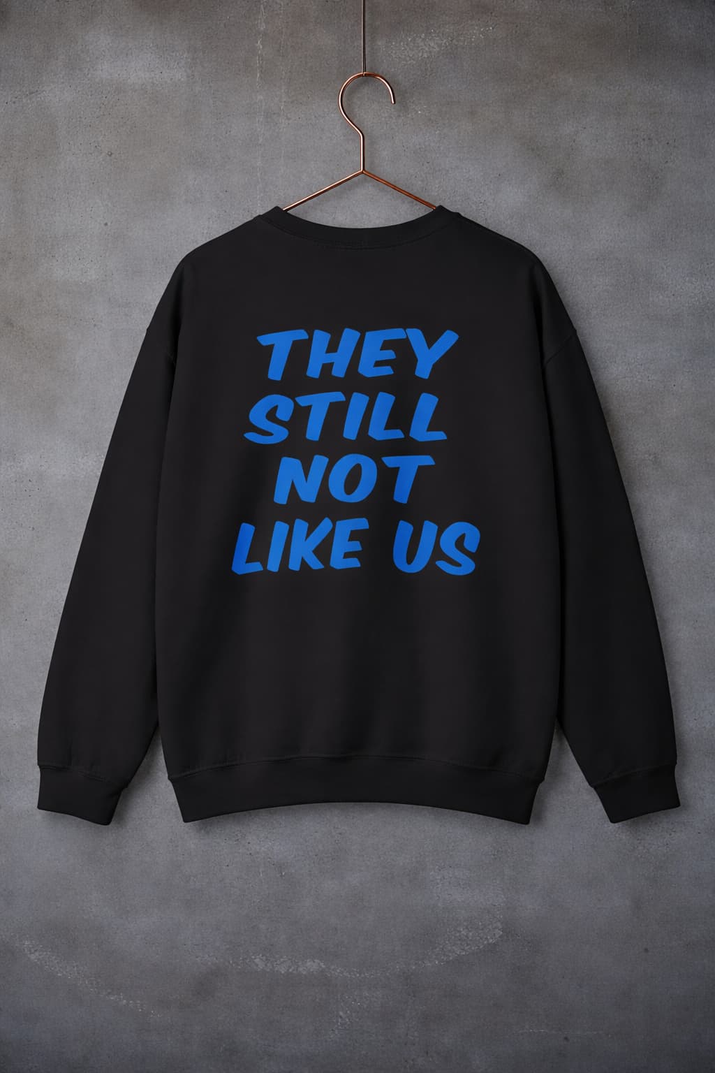 Sand color they still not like us crewneck sweatshirt hanging on copper hanger cement wall background