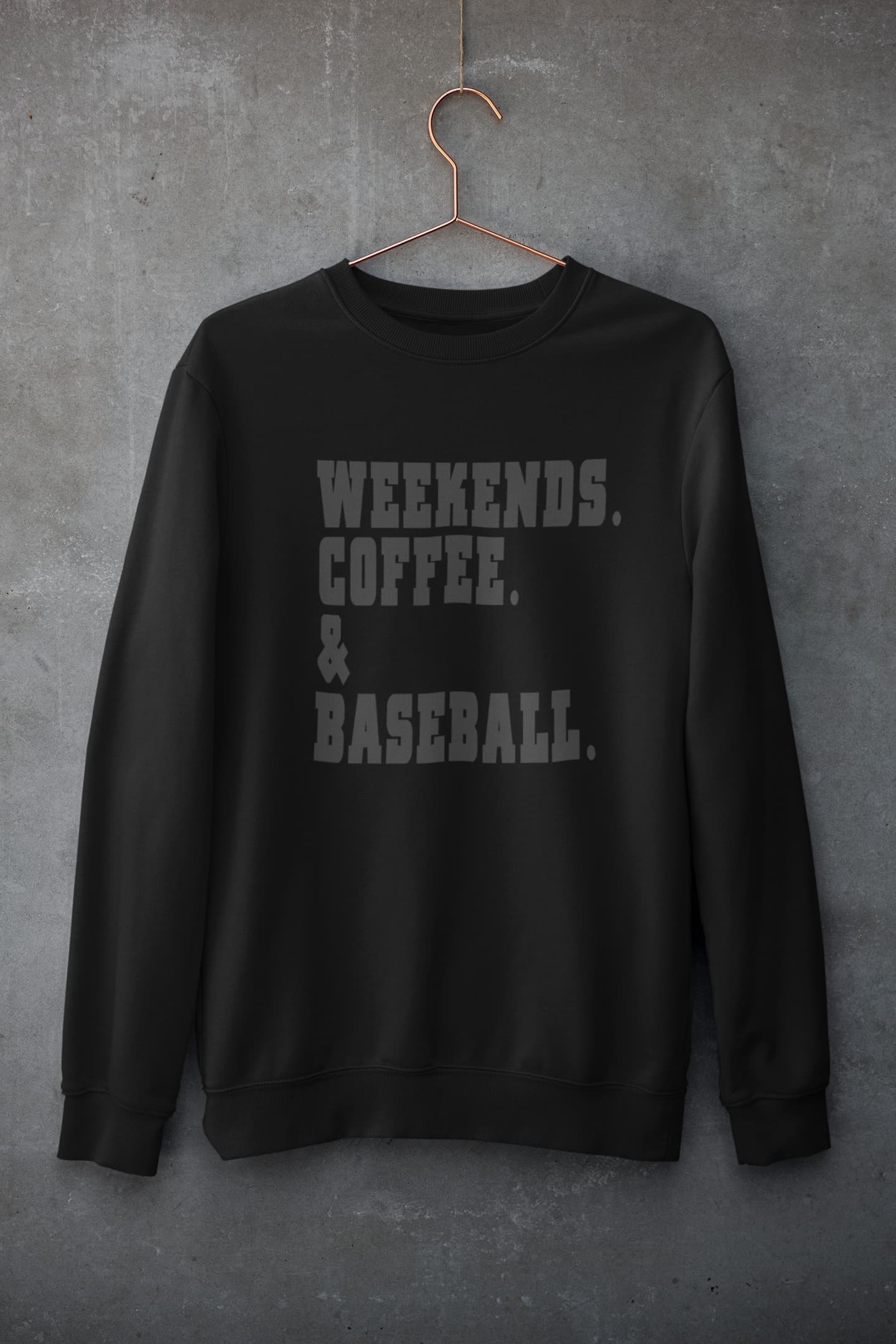Black weekends coffee baseball sweatshirt with bold varsity lettering hanging on hanger against concrete wall