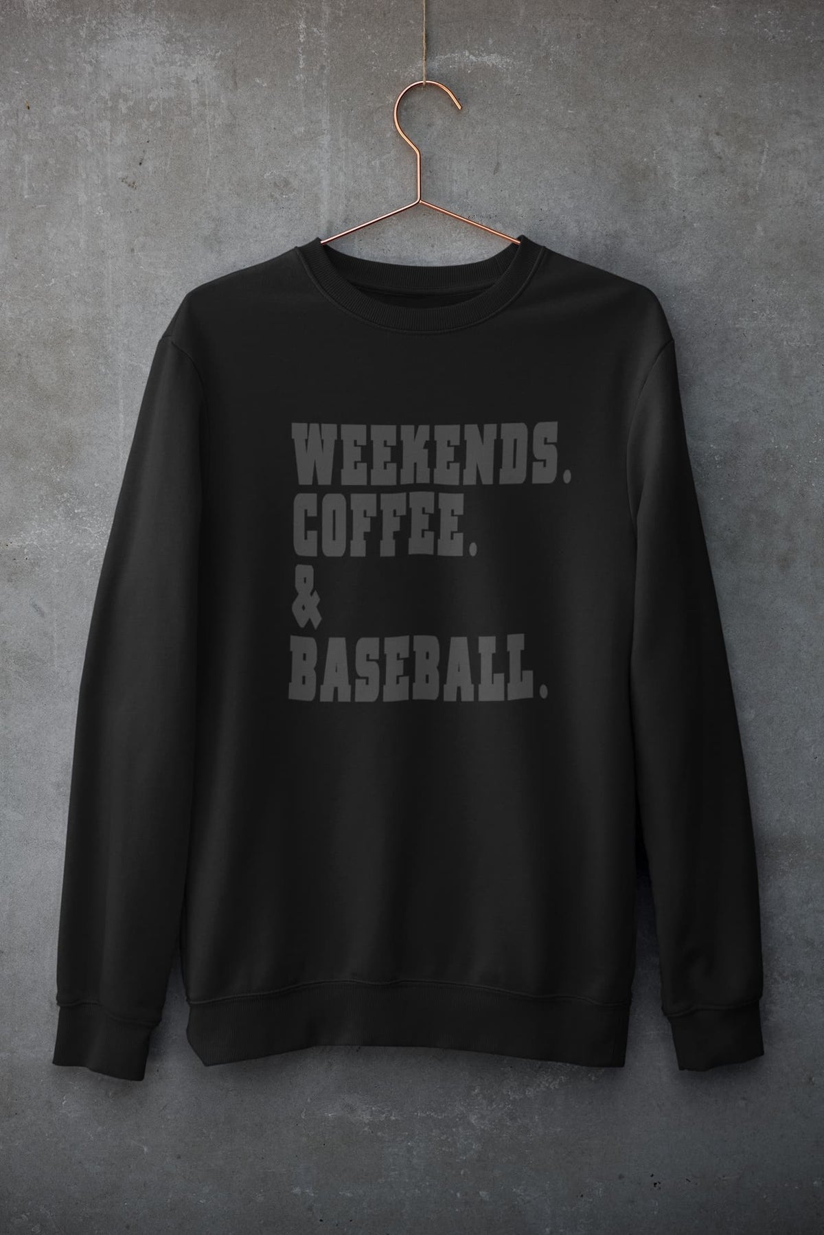 Black weekends coffee baseball sweatshirt with bold varsity lettering hanging on hanger against concrete wall
